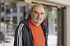 Vor einem Gebäude er tägt ein rotes T-Shirt und eine schwarze Trainingsjacke und schaut in die Kamera