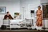 Krankenhauszimmer. Jemand liegt am Bett, an seiner Seite sitzt ein Mann, am offenen Fenster steht eine Frau mit Zigarette in der Hand