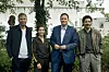 Die vier Personen stehen draußen vor Büchen und einem Baum, im Hintergrund Produktionsautos