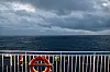 Reling bei Nacht mit Rettungsring, dahinter dunkel das Meer und Wolken