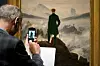 Ein Mann steht vor dem Gemälde, man sieht ihn von hinten, er fotografiert das Bild mit seinem Smartphone