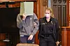 Zu Prozessbeginn wird der Angeklagte von einer Justizbeamtin in den Gerichtssaal geführt. er verdeckt sein Gesicht mit einem Aktenordner.