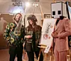 Benny Adrion und Kuratorin Hera stehen neben einer Staffelei mit einem Kunstwerk von Hera, rechts daneben steht ein Mensch im Anzug, der einen Karton in Form des Gallery-Logos auf dem Kopf trägt