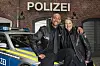Zwei Schauspieler lehnen an einem Polizeiwagen.