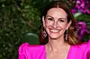 Schauspielerin Julia Roberts in einem pinken Kleid mit großen Uhrringen, lachend, hinter ihr ist eine Wand aus Pflanzen.