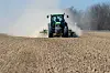 Traktor auf einem Feld wirbelt Staub auf, im Hintergrund stehen kahle Bäume.