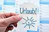 Eine Hand hält einen Zettel mit der Aufschrift „Urlaub“ vor einem Kalender.