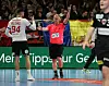 Der Schiedsrichter hebt den linken Arm und streckt zwei Finger nach oben. Den rechten Arm hält er gerade vor den Körper weg, auf den Spieler gerichtet.