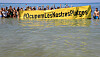 Viele Menschen stehen am Strand und halten ein Plakat hoch, auf dem steht ”Besetzen wir (selbst) unsere Strände“