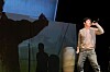 Darsteller auf der Bühne, einige im Schatten, einer - mit Mikrofon in der Hand - im Scheinwerferlicht