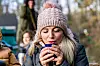 Eine Frau mit blonden Haaren und Bommelmütze wärmt sich die Hände an einer Tasse Glühwein.