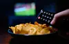 Ein Junge hält eine Fernbedienung eines Fernsehers neben einer Schüssel mit Kartoffelchips in der Hand. (Symbolbild)