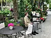 Person sitzt in einem blumengeschmückten Straßencafé an einem Tisch mit Getränken.