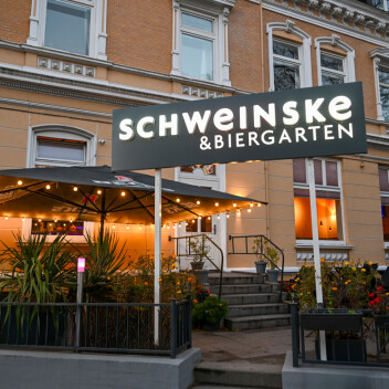 Schweinske-Restaurant mit Biergarten und Lichterketten an einer Straßenecke am Abend.