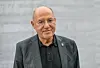 Gregor Gysi trägt ein dunkles Sakko und ein schwarzes Poloshirt, blickt frontal in die Kamera und steht vor einem hellgrauen Hintergrund.