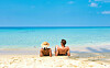 Mann und Frau sitzen nebeneinander an einem weißen Sandstrand und blicken auf das türkisfarbene Meer. Im Vordergrund liegen Strand und Wasser unter blauem Himmel.