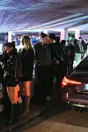 Mehrere junge Frauen in Overknees, Blazern und dunkler Streetwear stehen neben Sportwagen in einem bunt beleuchteten Parkhaus.