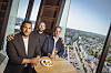 Restaurantleiter Ramzi Rahali, Küchenchef Maurice Exner und Bardirektor Bastian Knülle stehen im „Clouds“ vor bodentiefen Fenstern mit Blick über Hamburg. Auf dem Tisch stehen eine Zitronentarte und ein Glas Schaumwein.
