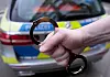 Eine Hand hält ein paar Handschellen. Im Hintergrund ist ein Polizeiauto zu sehen.