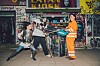 Drei Schauspieler inszenieren einen Kampf vor einem Kiosk.