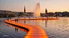 Wasserweg (Binnenalster): Auf der Binnenalster führt ein leuchtender Steg über das Wasser und eröffnet neue Blicke auf die Innenstadt. Leichtes Schaukeln und die Brise der Alsterfontäne machen den Weg zu einem besonderen Erlebnis für alle. (Visualisierung KI unterstützt)