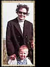 Ein kleiner Junge steht vor seiner Mutter, die eine schwarze Jacke und eine Sonnenbrille trägt.