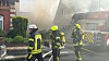 Feuerwehrkräfte löschen einen Brand vor einem Wohnhaus, dichter Rauch steigt auf.