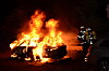 Ein brennendes Auto in der Nacht, daneben Einsatzkräfte in Schutzkleidung.
