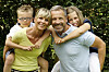 Familie im Garten: Vater, Mutter und zwei Kinder lachen und umarmen sich vor grünem Hintergrund.