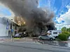Dichter Rauch über einem brennenden Gebäude, Feuerwehrfahrzeuge und Einsatzkräfte an der Straße.