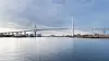 Große Brücke über ruhigem Wasser, dahinter Ufer mit Gebäuden und klarer Himmel.