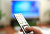 Mann drückt Knopf auf der Fernbedienung vor einem unscharfen Fernseher.