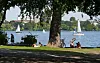 Menschen entspannen im Schatten an der Außenalster, Segelboote auf dem Wasser.
