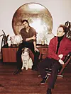 Zwei Männer sitzen in einem Retro-Wohnraum, ein Hund steht zwischen ihnen, großer Rundspiegel im Hintergrund.