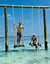 Alexander Zverev und Sophia Thomalla schaukeln an Seilen über seichtem, türkisfarbenem Wasser vor blauem Himmel.
