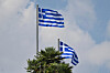 Zwei griechische Nationalflaggen wehen vor bewölktem Himmel über Palmen.