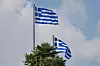 Zwei griechische Nationalflaggen wehen vor bewölktem Himmel über Palmen.