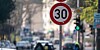 Tempo-30-Schild an einer belebten Stadtstraße mit Autos und Ampel im Hintergrund.