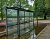 Verlassene verglaste Haltestelle in einem Park, umgeben von Wasser und Bäumen.