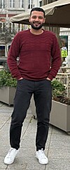 Mann steht auf dem Gehweg vor einem Café mit Sonnenschirm und Sitzbereich.
