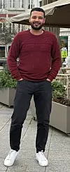 Mann steht auf dem Gehweg vor einem Café mit Sonnenschirm und Sitzbereich.