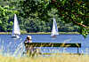 Frau auf Gartenbank beobachtet zwei Segelboote auf der Havel bei Sonne.