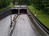 Nasse Zufahrtsstraße mit Absperrungen und Warnlichtern vor einem zweigeteilten Tunnel in grüner Umgebung.
