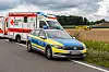 Rettungswagen und Polizeiauto stehen an einer Landstraße neben einem Feld, im Hintergrund ein gelbes Einsatzfahrzeug.