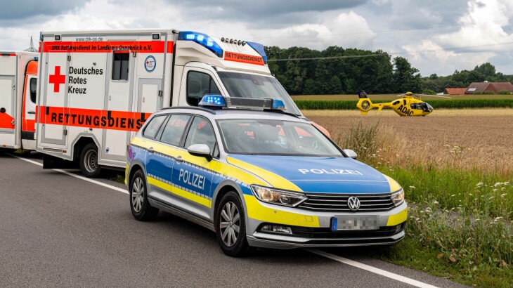 Rettungswagen und Polizeiauto stehen an einer Landstraße neben einem Feld, im Hintergrund ein gelbes Einsatzfahrzeug.