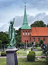 Statue im Friedhof vor Backsteinkirche mit grünem Turm und rotem Dach.