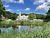 Weißes Herrenhaus hinter Teich, umgeben von Bäumen, Schilf und blauem Himmel mit Wolken.