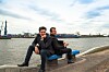 Zwei Männer sitzen auf einer blauen Bank am Hafen mit Industrieanlagen im Hintergrund.