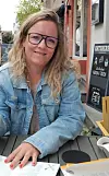 Frau mit Brille sitzt an einem Tisch im Straßencafé und lächelt in die Kamera.