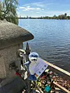Teddybär mit Brille hält ein Blatt am Ufergeländer vor einem See.